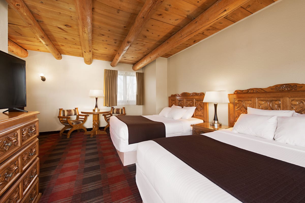 A hotel room with two beds, a wooden ceiling, a television, a desk with a lamp, and a plaid carpet.
