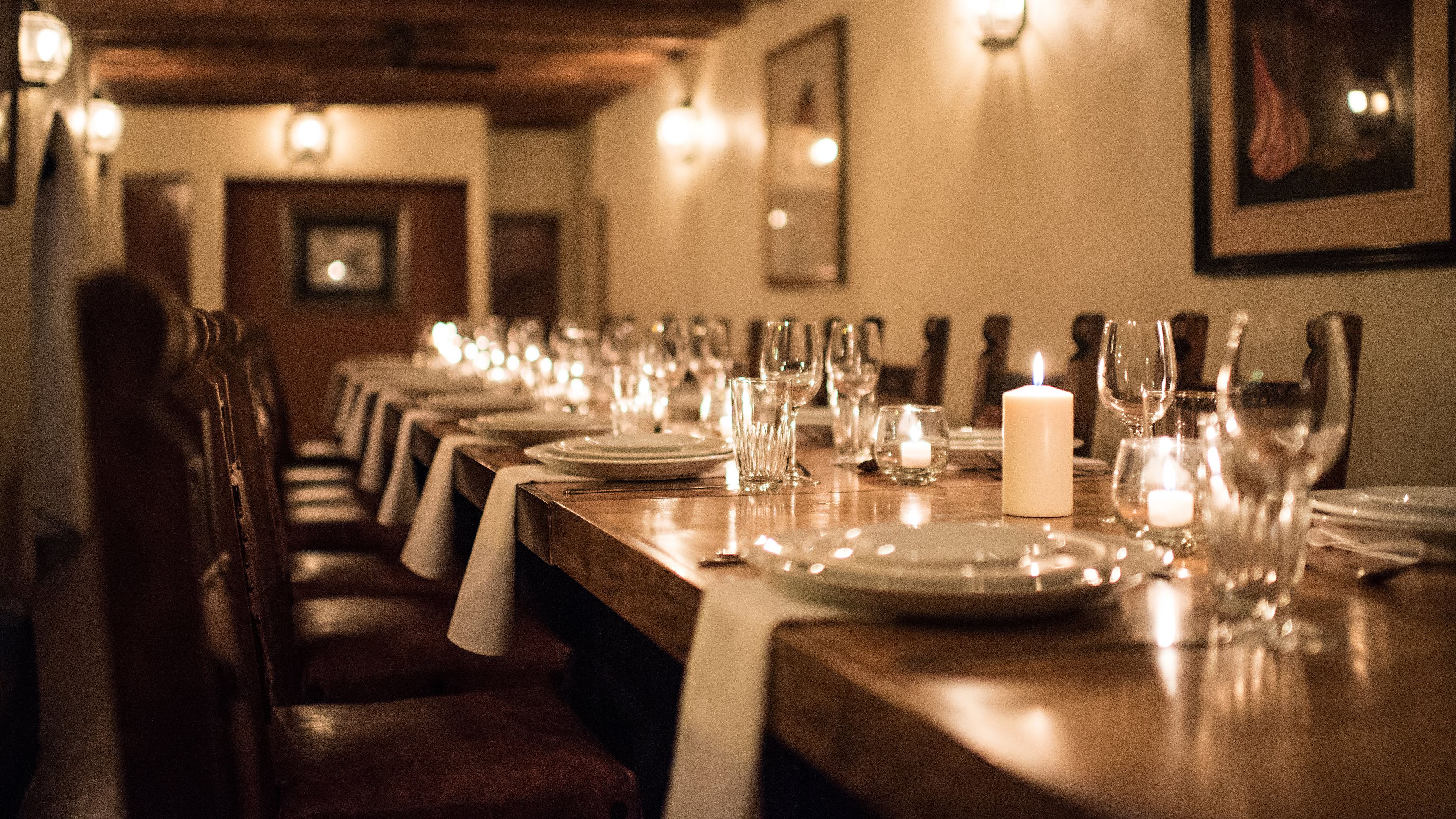 Ein Restaurant mit einem langen Holztisch, gedeckt für eine Mahlzeit, mit Tellern, Gläsern und einer brennenden Kerze.