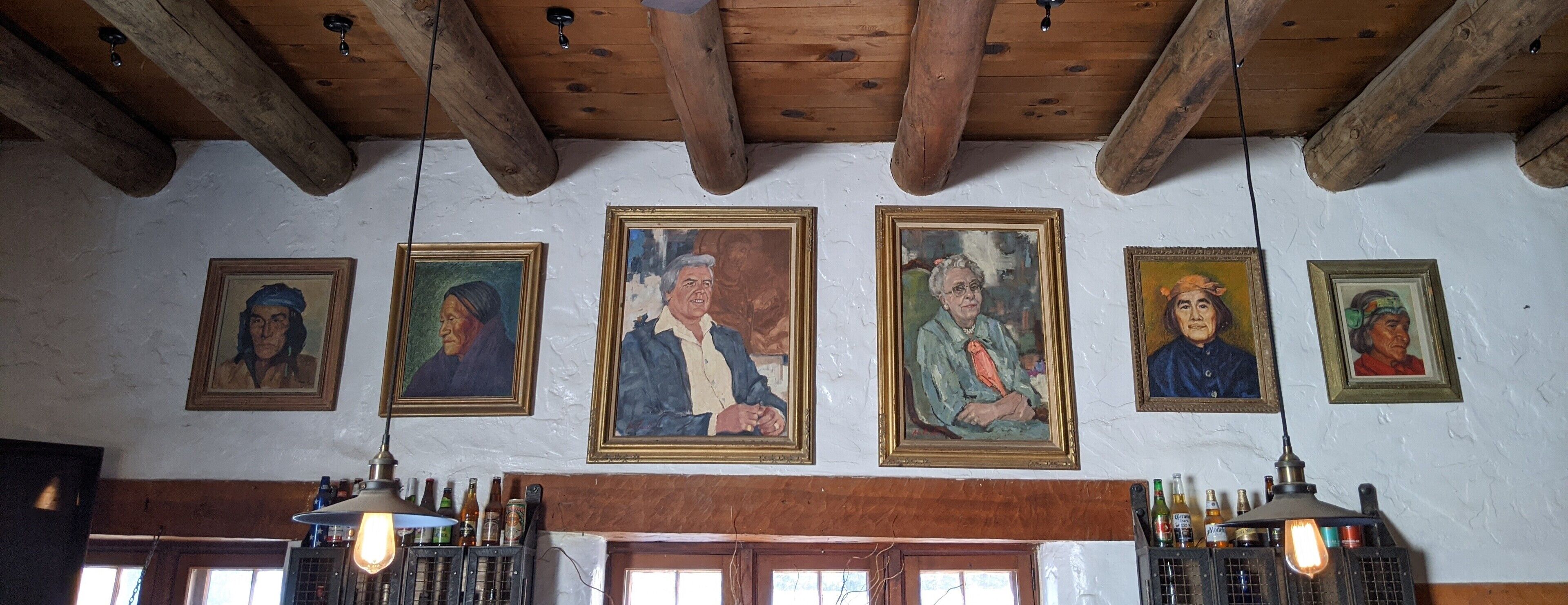 A room with wooden beams on the ceiling, a white wall with framed portraits, and a bar area with bottles and a light fixture.