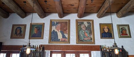 A room with wooden beams on the ceiling, a white wall with framed portraits, and a bar area with bottles and a light fixture.
