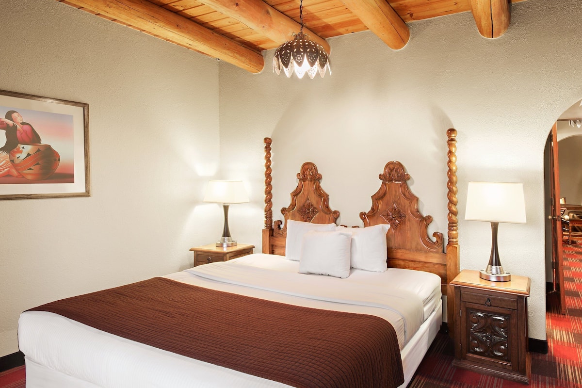 A hotel room with a wooden ceiling, a large bed with a brown headboard, two bedside tables with lamps, and a painting on the wall.