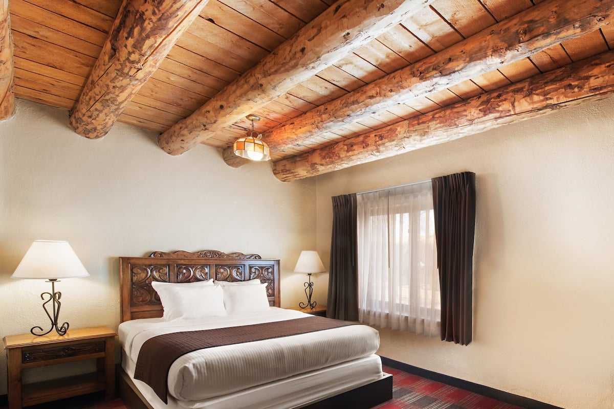 A hotel room with a wooden ceiling, a bed with a headboard, two bedside tables with lamps, a window with curtains, and a checkered carpet.