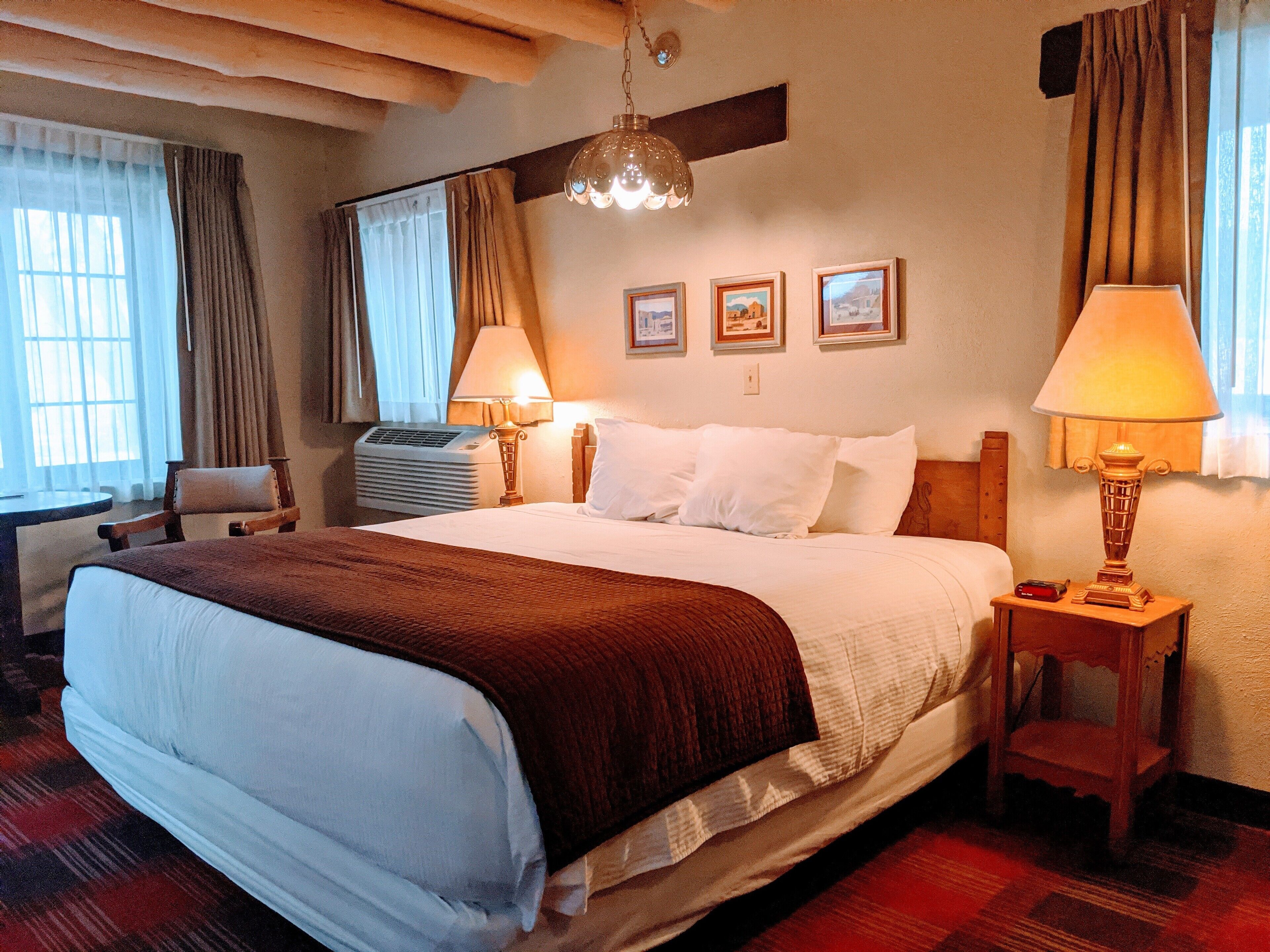 A hotel room with a large bed, two bedside lamps, a wooden headboard, a small table, a chair, and a window with curtains.