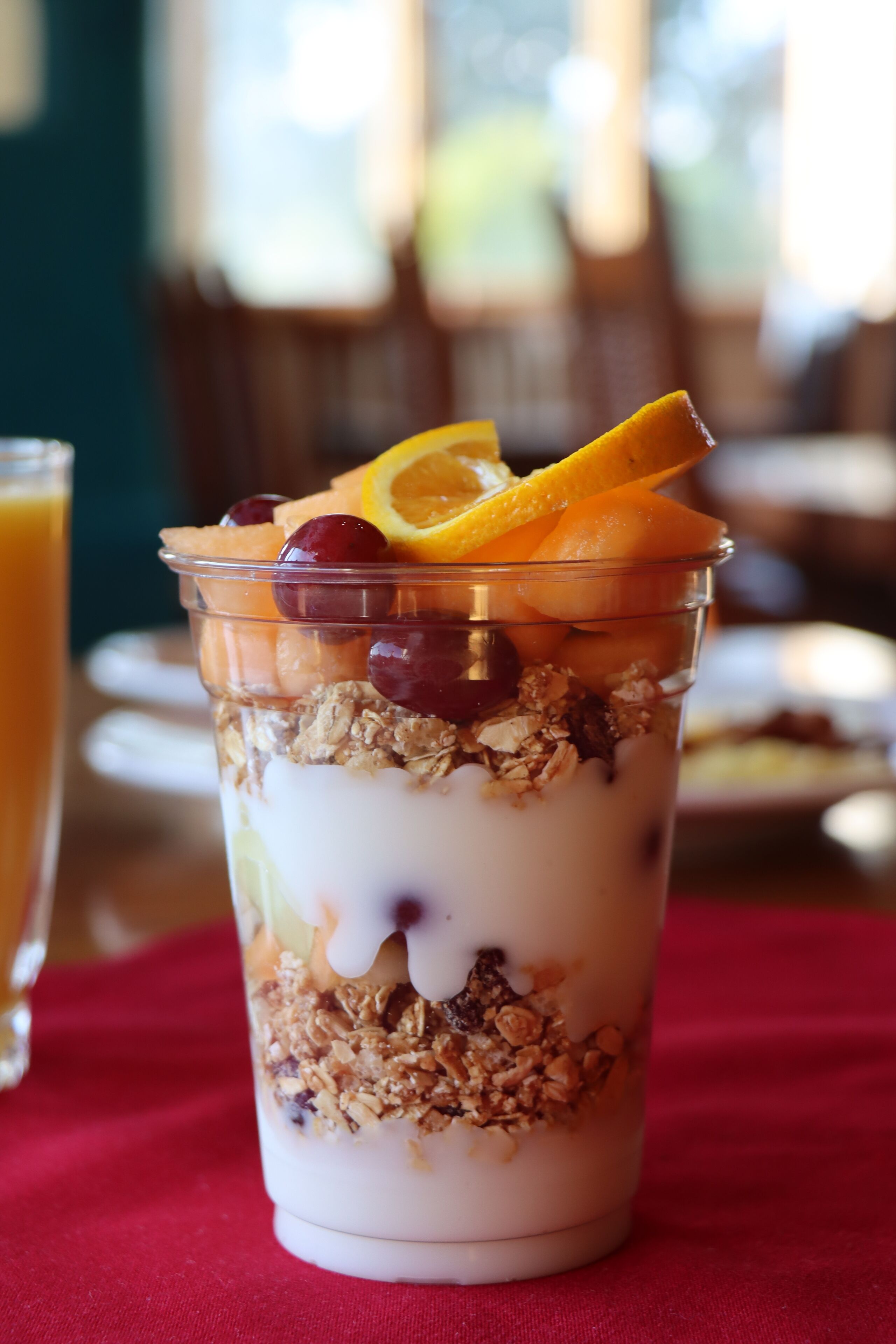 A layered dessert in a clear cup with fruits and granola on a red tablecloth.