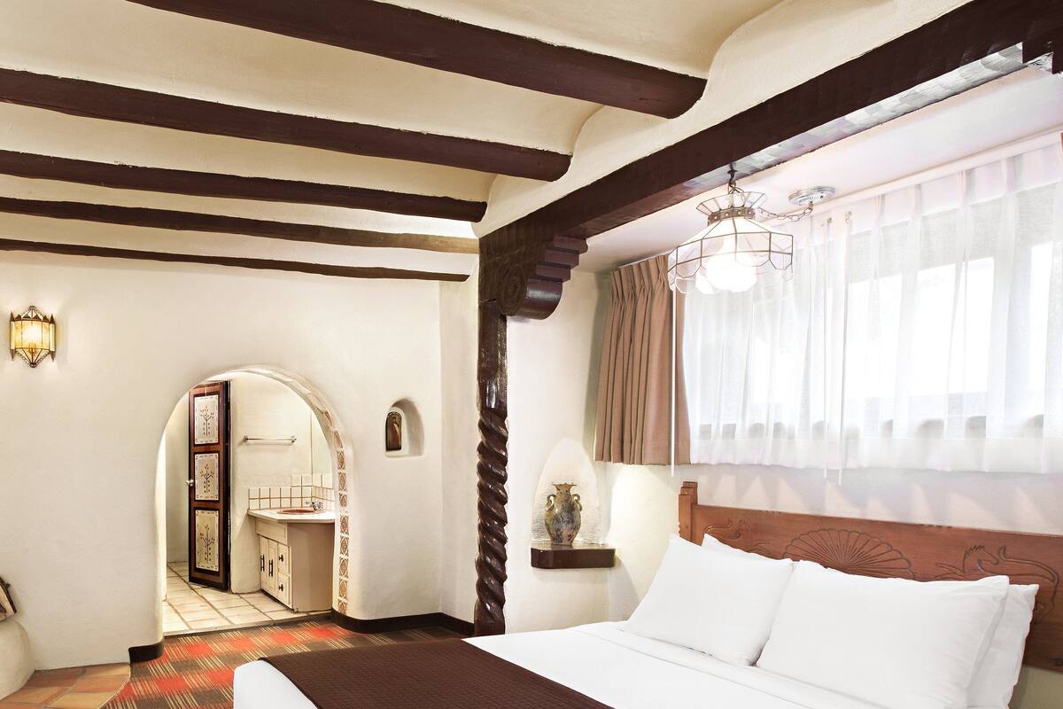 A hotel room with a large bed, a wooden headboard, a window with sheer curtains, a small table with a lamp, and a bathroom visible through an arched doorway.