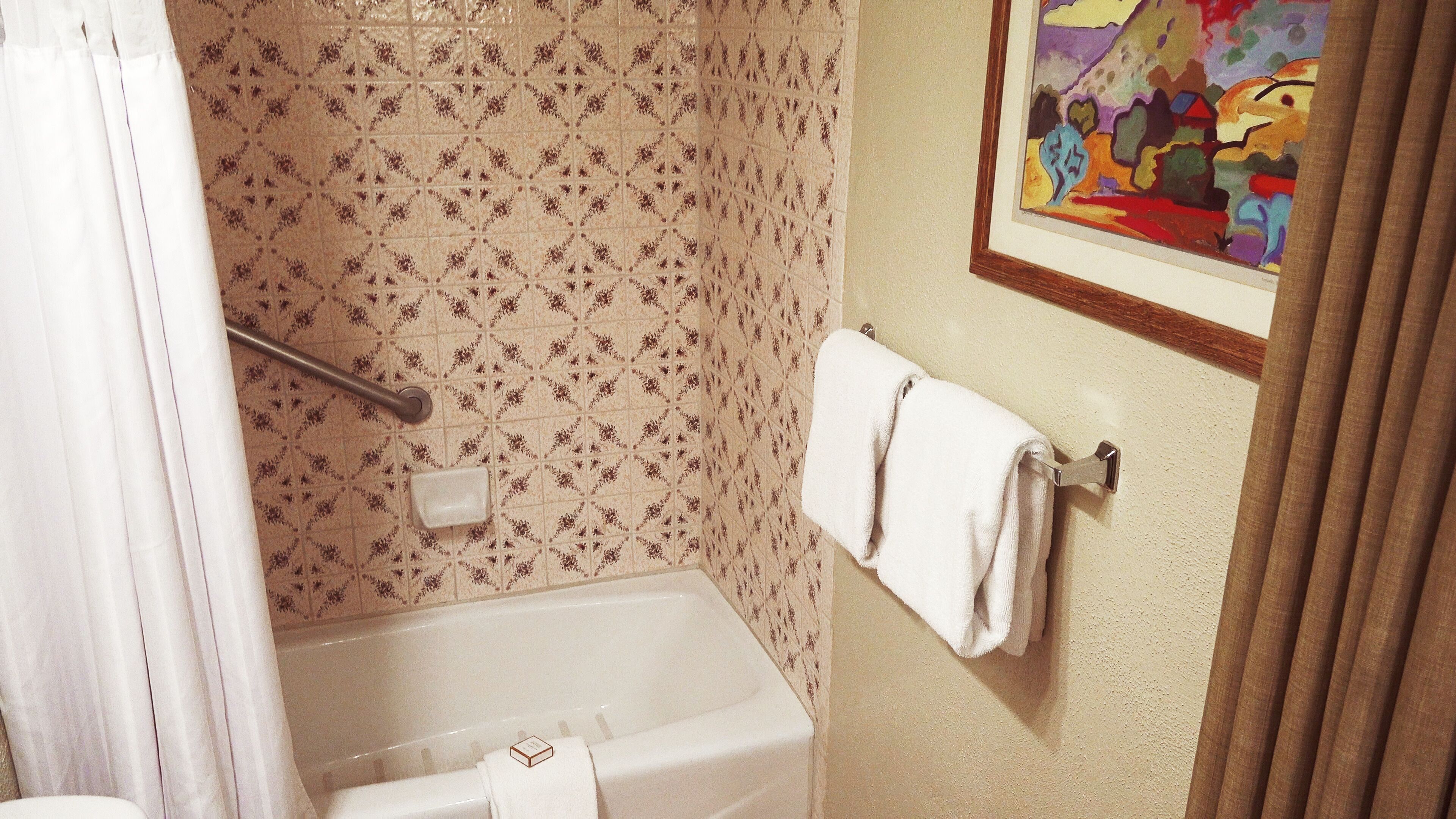 A bathroom with a bathtub, a shower curtain, a towel rack with a towel, and a colorful painting on the wall.