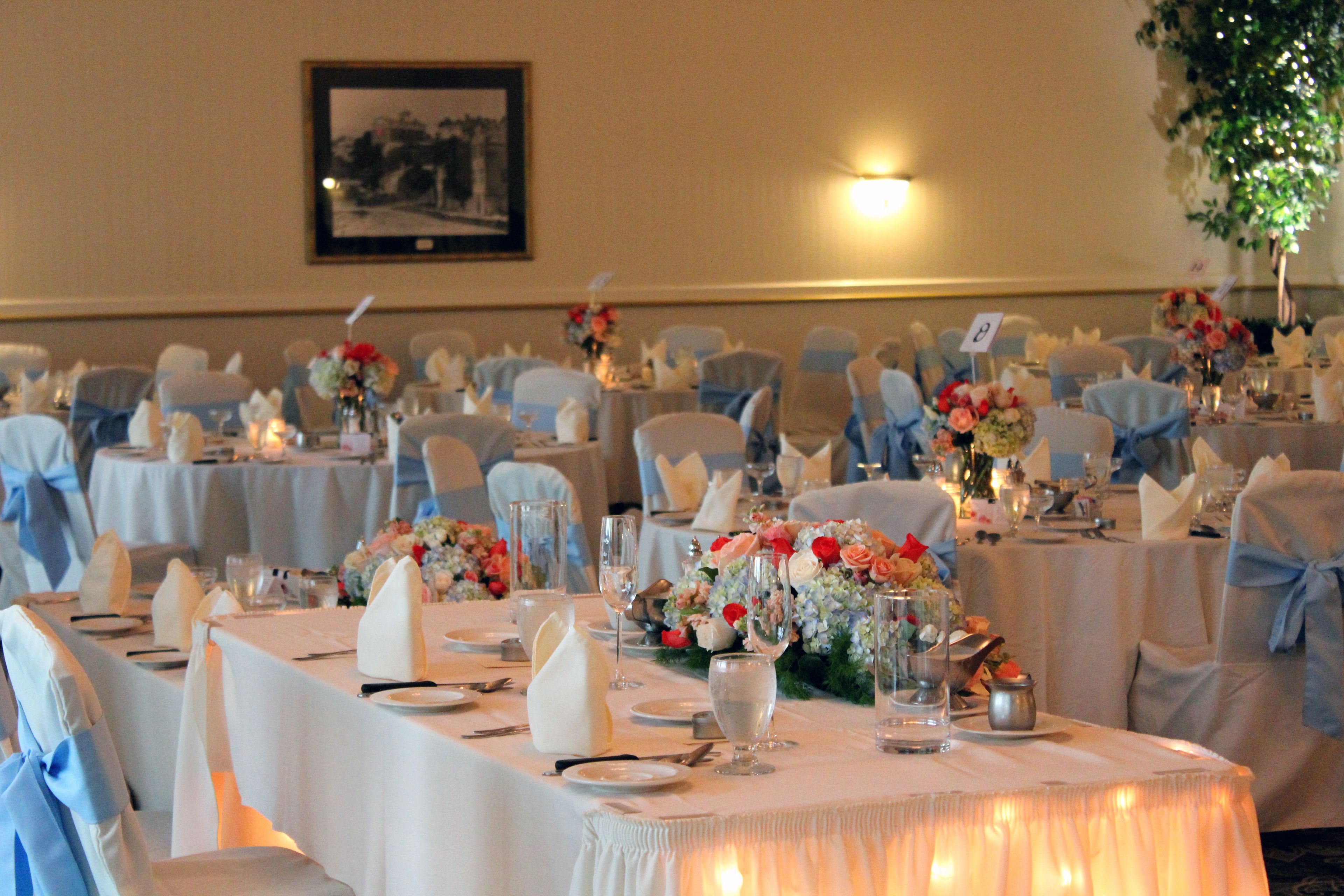 A banquet hall with round tables set for a formal event, featuring white tablecloths, blue chair covers, and floral centerpieces.