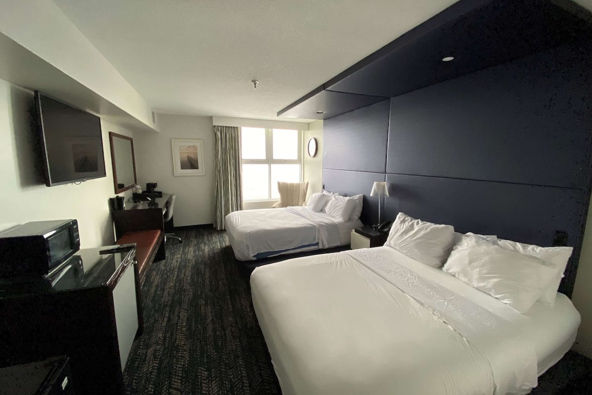 Ein Hotelzimmer mit zwei Betten, einem Schreibtisch mit Fernseher, einer Uhr und einem Fenster mit Vorhängen.