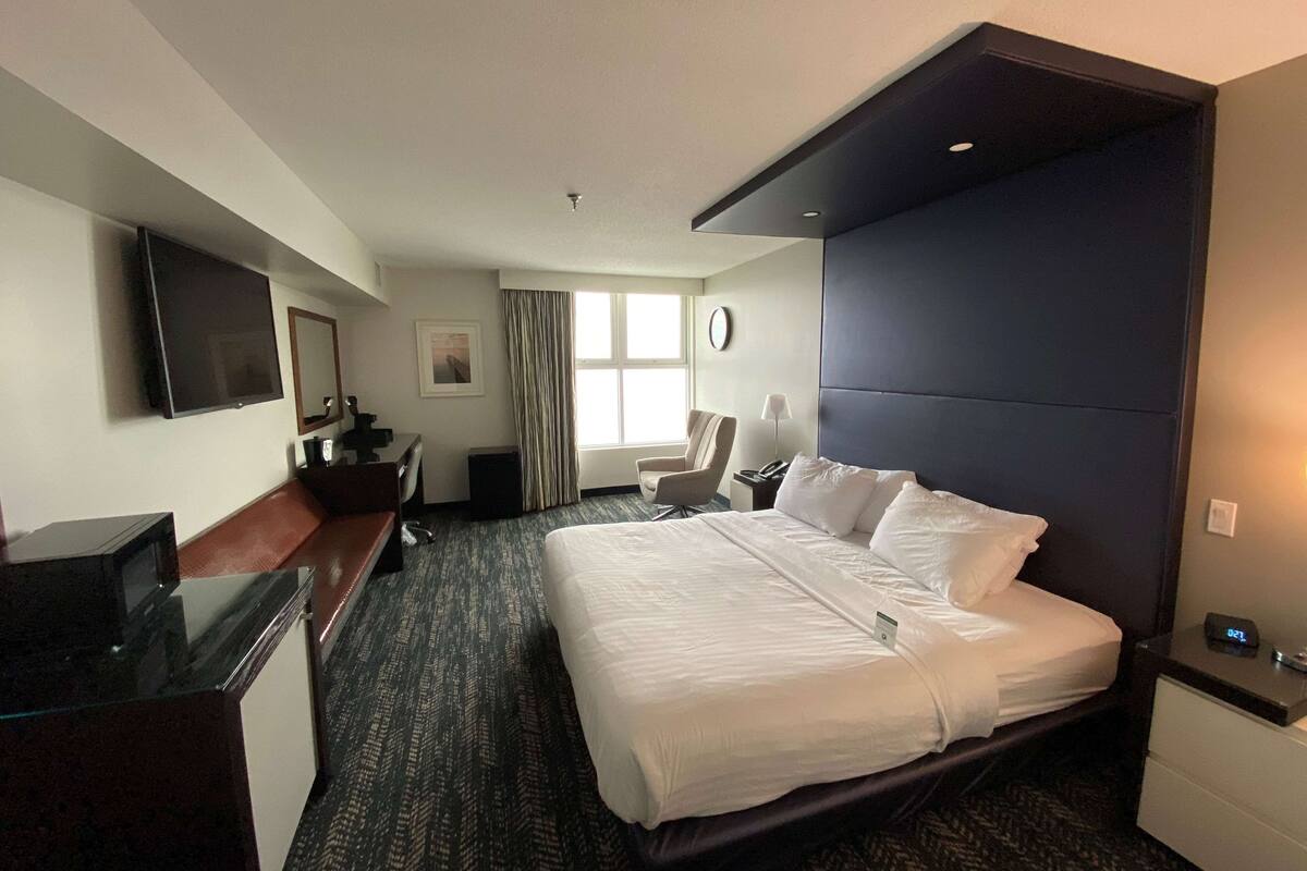 Ein Hotelzimmer mit einem großen Bett, einem Schreibtisch mit Computer, einem an der Wand befestigten Fernseher, einem Sessel und einem Fenster mit Vorhängen.