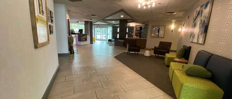 Een hotel lobby met een visgraat vloerpatroon, een receptiebalie, een zithoek met groen en bruin gestoffeerde stoelen, en een groot abstract schilderij aan de muur.