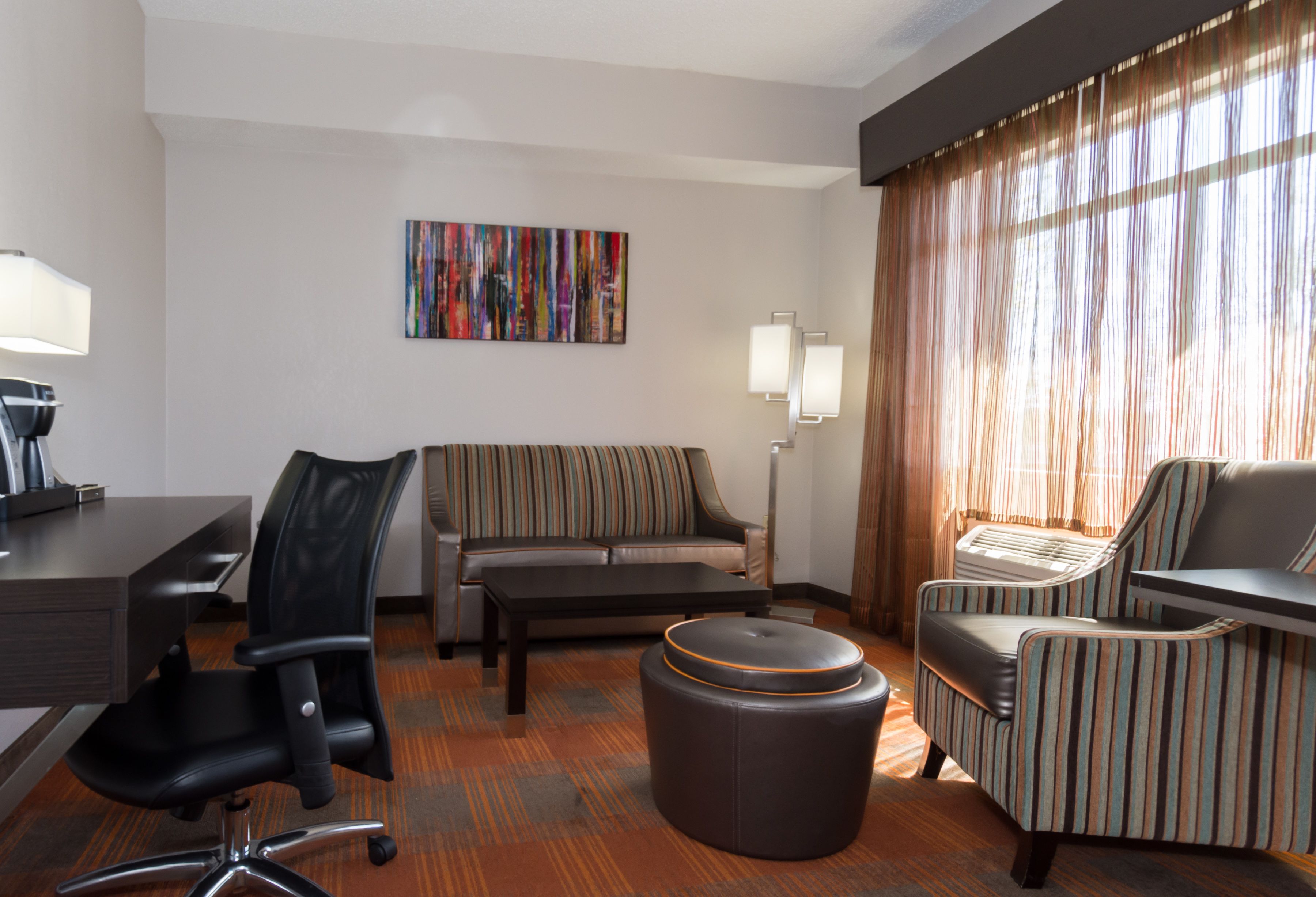 Habitación de hotel con escritorio, silla de oficina, sofá, otoman, sillón a rayas y un cuadro en la pared.