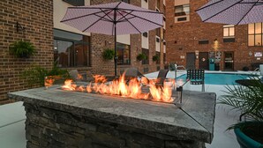 Un foyer en pierre avec des flammes, des sièges d’extérieur et des parasols, le tout situé près d’une piscine dans un bâtiment en briques.