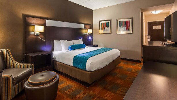 Une chambre d’hôtel avec un grand lit, deux lampes de chevet, une chaise, un petit pouf et un bureau avec un téléviseur à écran plat.