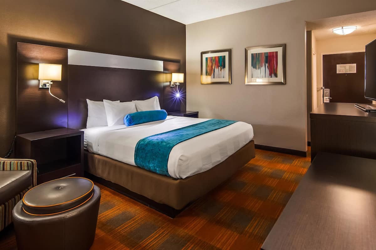Een hotelkamer met een groot bed, twee bedlampjes, een stoel, een klein poufje en een bureau met een flatscreen televisie.