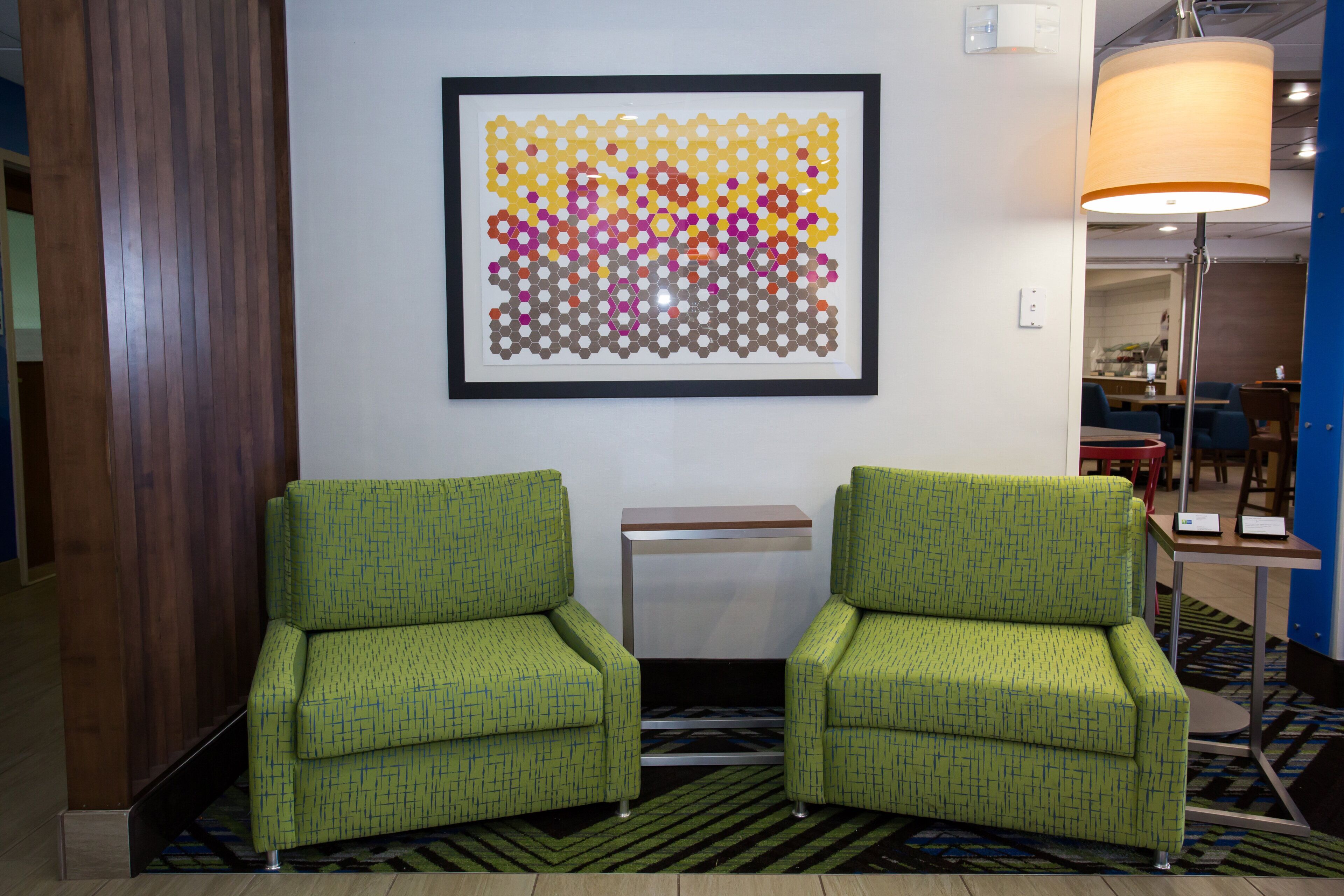 A hotel lobby with two green armchairs, a framed abstract art piece, a small table, and a floor lamp.