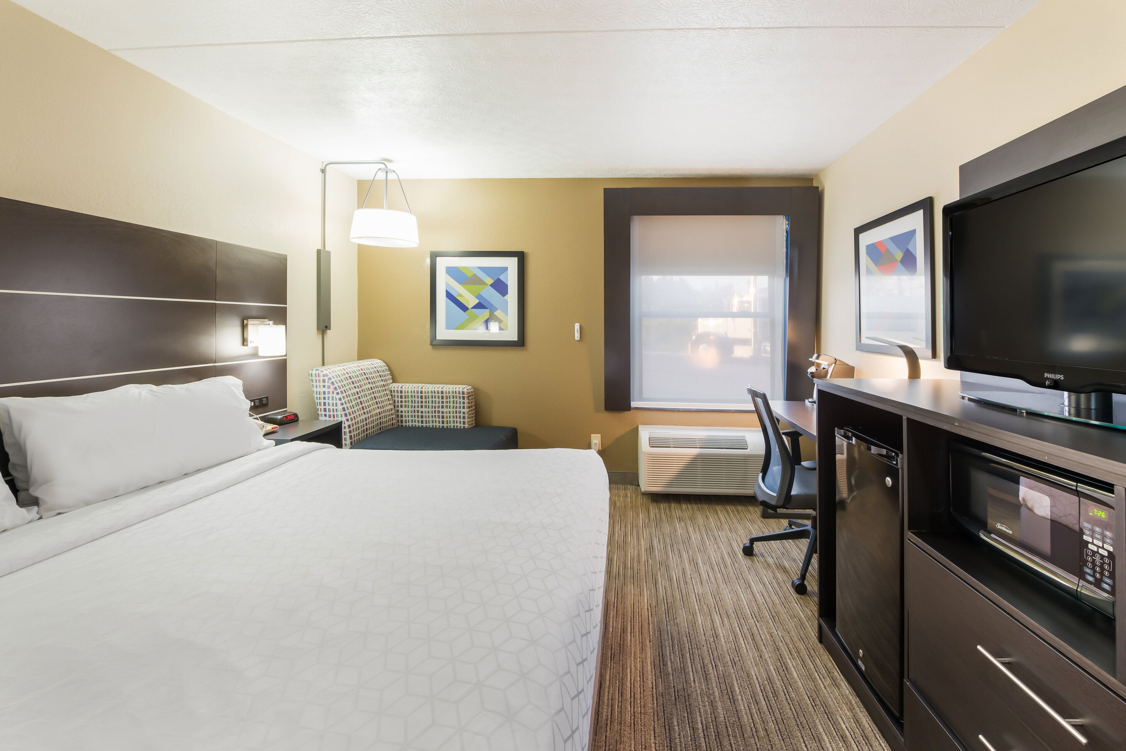 A hotel room with a large bed, a desk with a flat-screen TV, a microwave, and a heater. There is a window with a view of the outside.