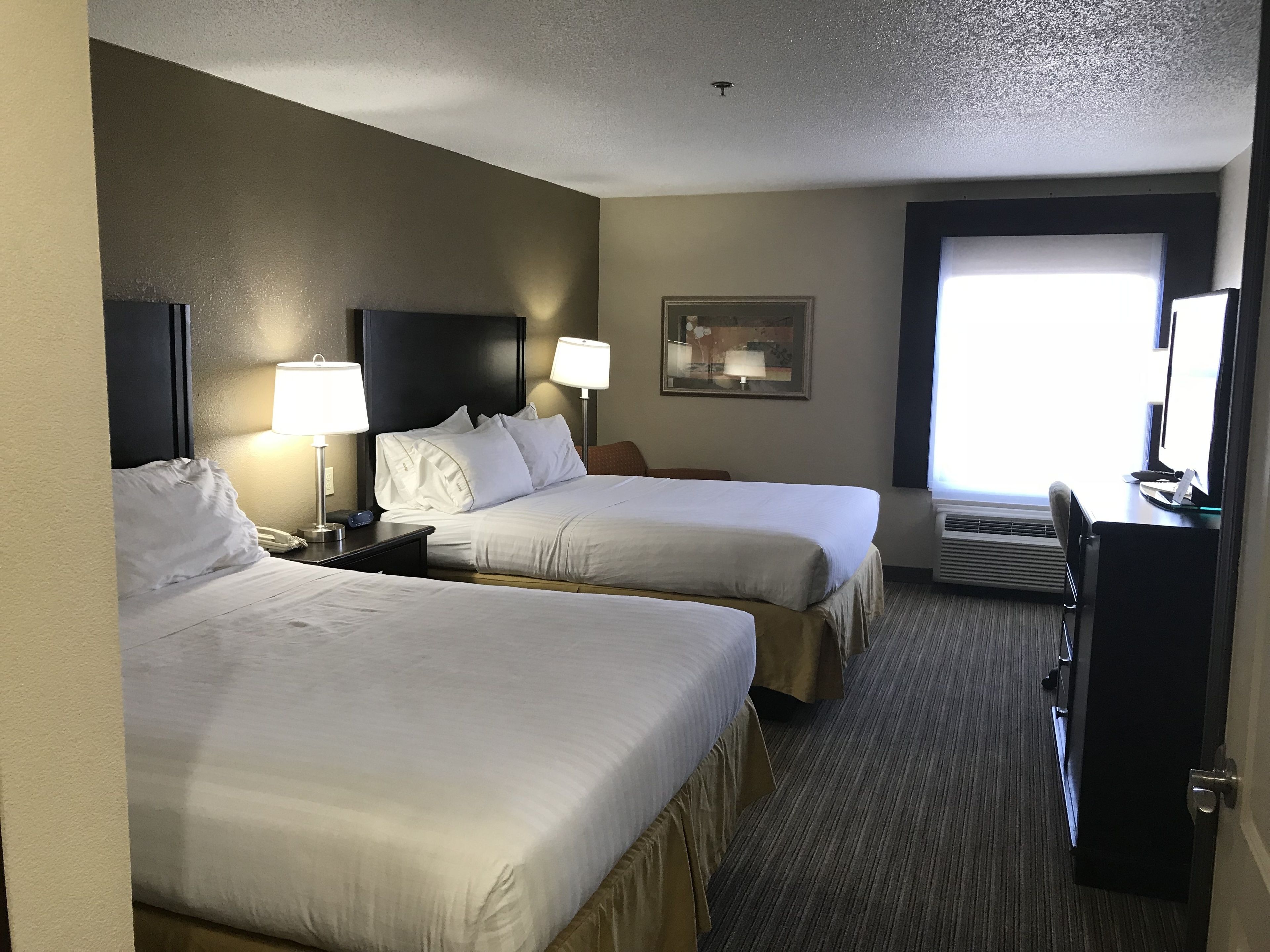 Une chambre d’hôtel avec deux lits, un bureau, une chaise, une lampe, un téléphone et une fenêtre avec des stores.