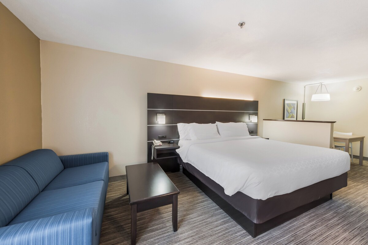 A hotel room with a large bed, a sofa, a bedside table, a nightstand, and a desk with a lamp.