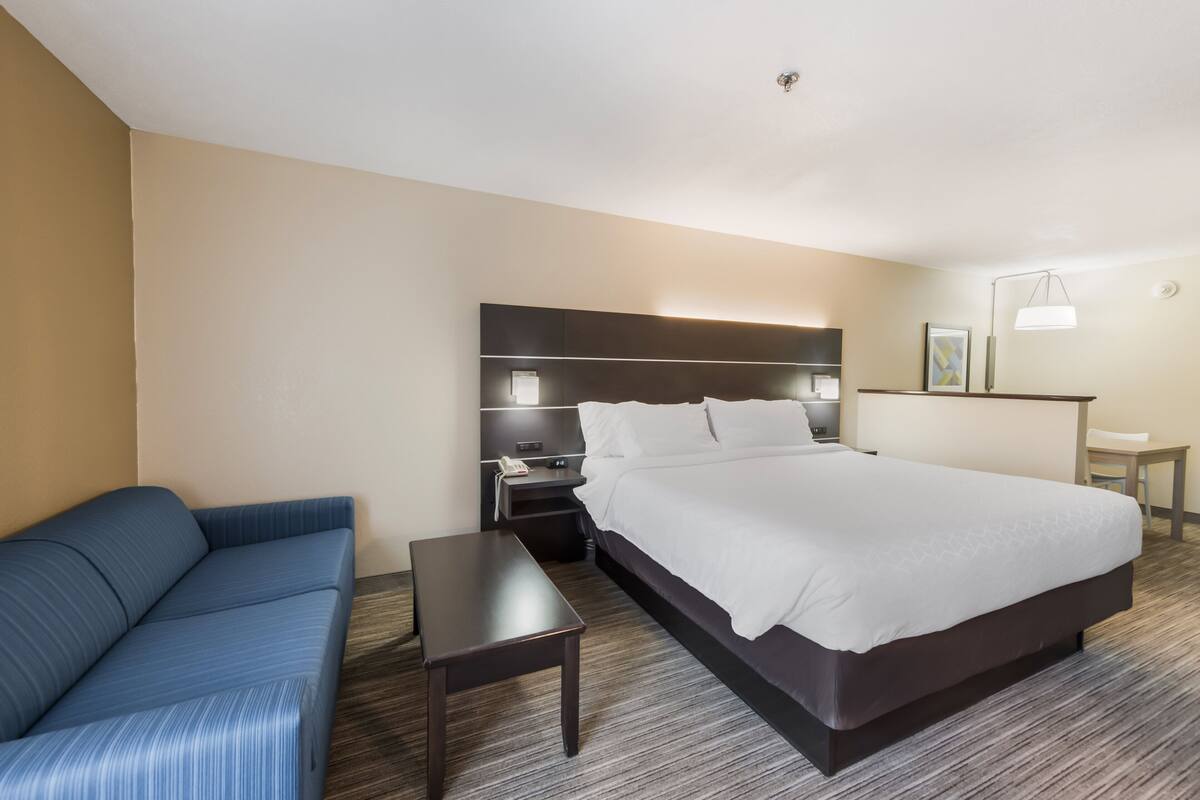 A hotel room with a large bed, a sofa, a bedside table, a nightstand, and a desk with a lamp.