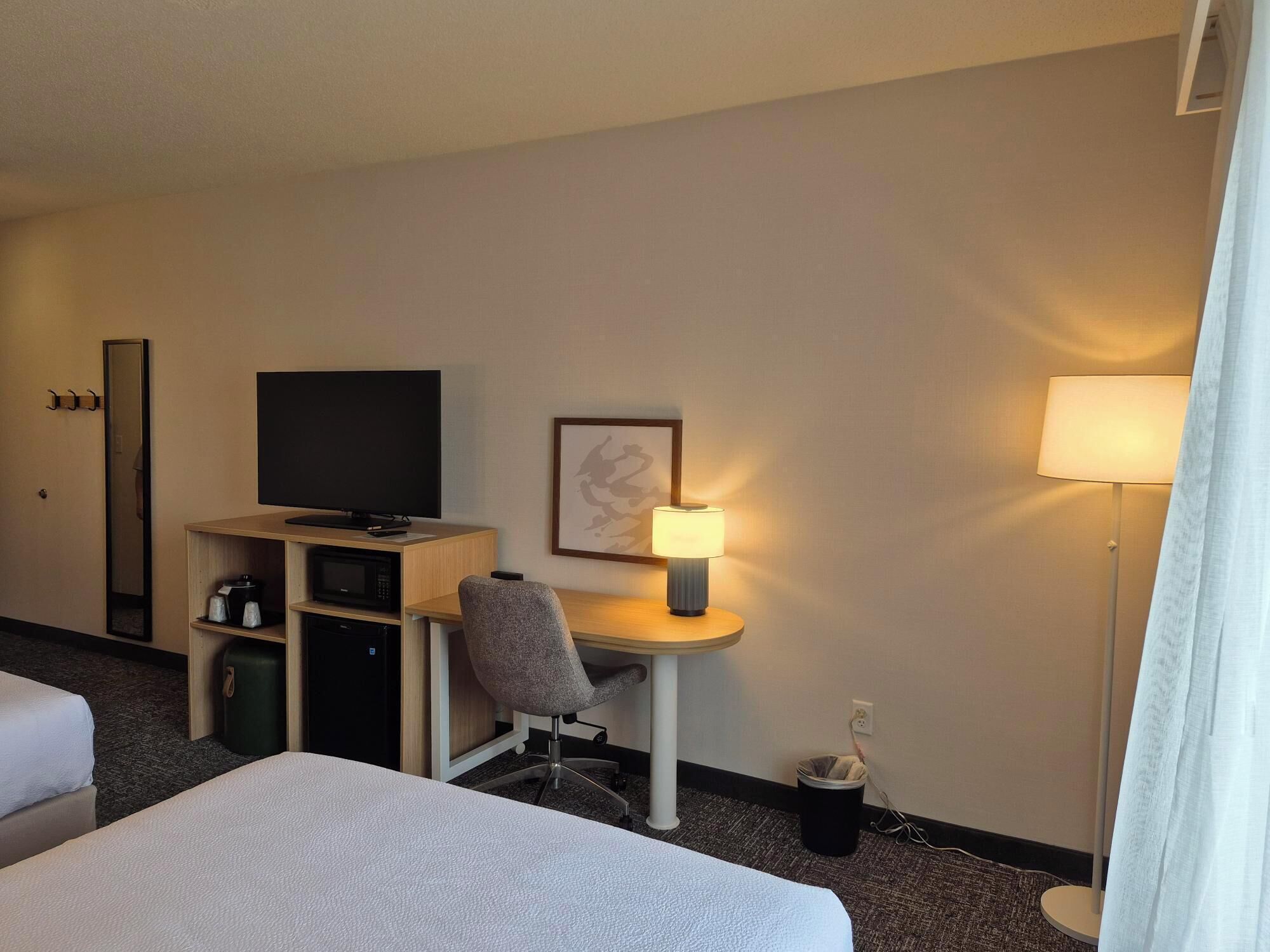 A hotel room with a bed, a desk with a chair, a television, a lamp, and a trash bin.