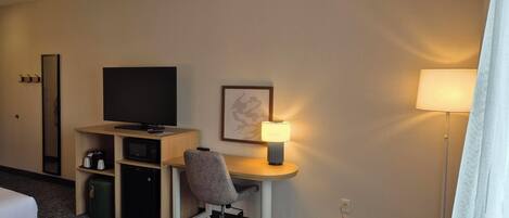 A hotel room with a bed, a desk with a chair, a television, a lamp, and a trash bin.