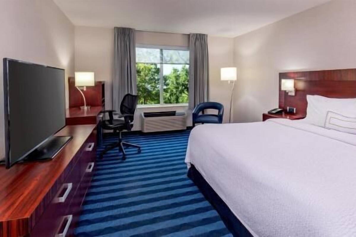 A hotel room with a bed, a desk with a chair, a television, and a window with curtains.