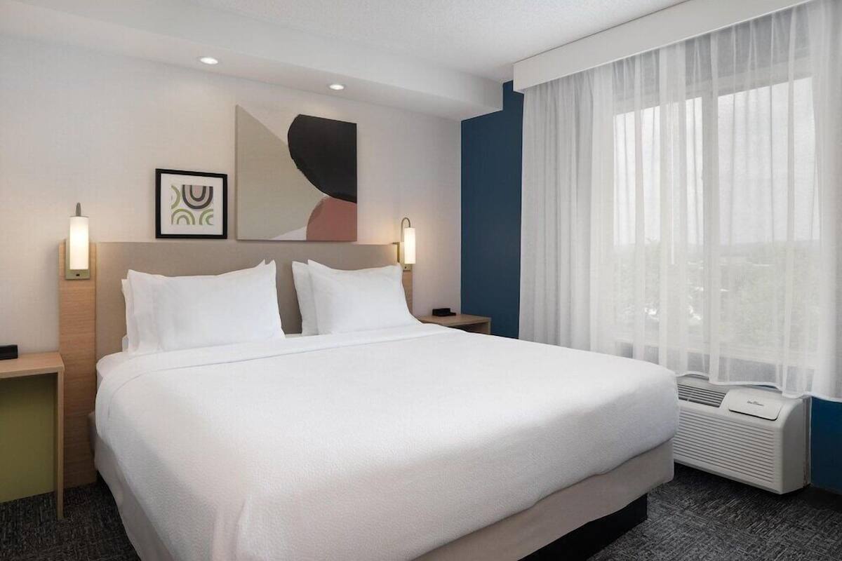 A hotel room with a large bed, bedside tables, a mirror, and a window with sheer curtains.
