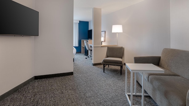A hotel room with a flat-screen TV mounted on the wall, a desk with a lamp, a grey sofa, and a chair. - Spark by Hilton Lancaster (Lancaster)