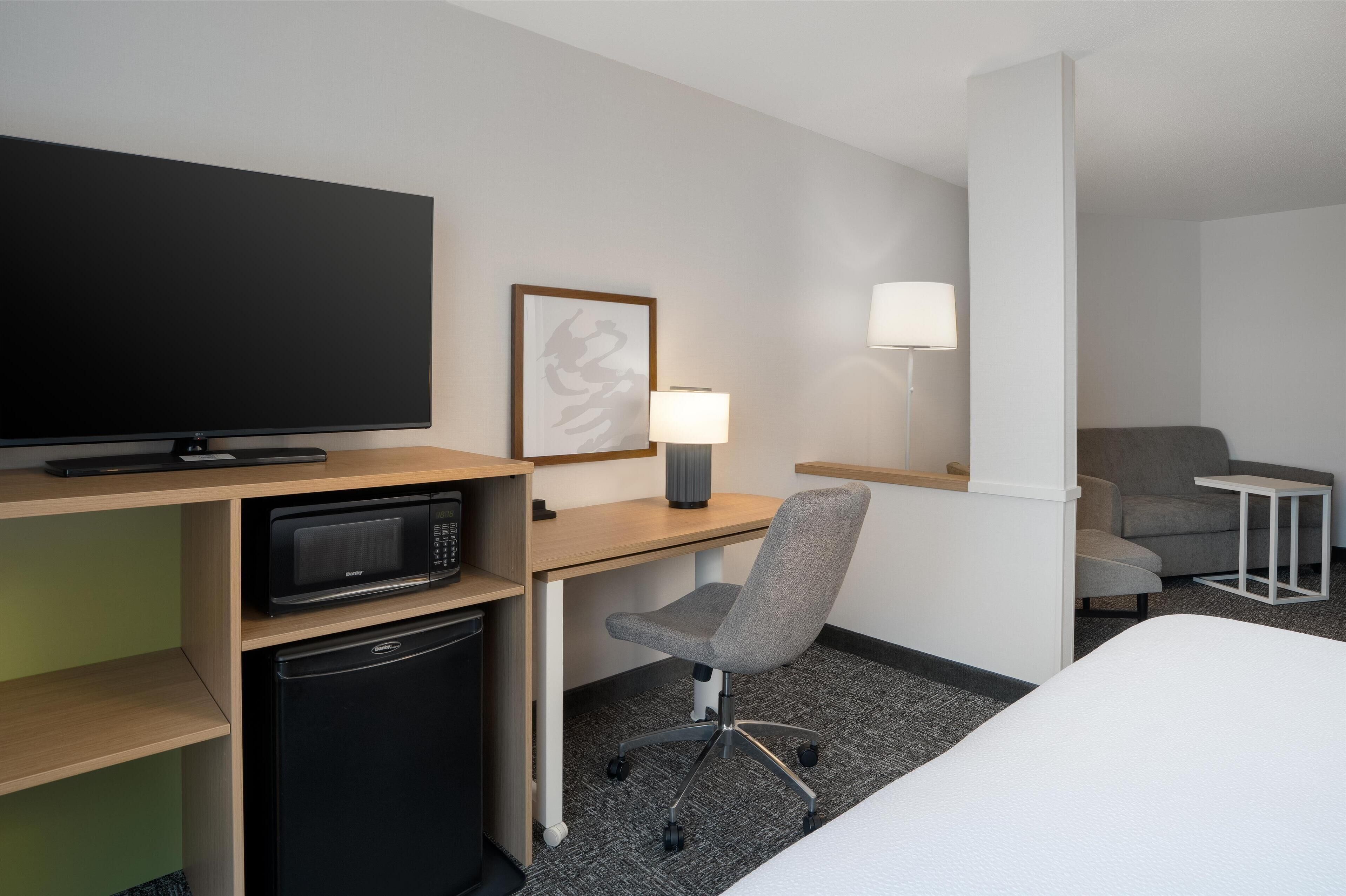 Et hotelværelse med seng, skrivebord med computer, mikroovn og køleskab.