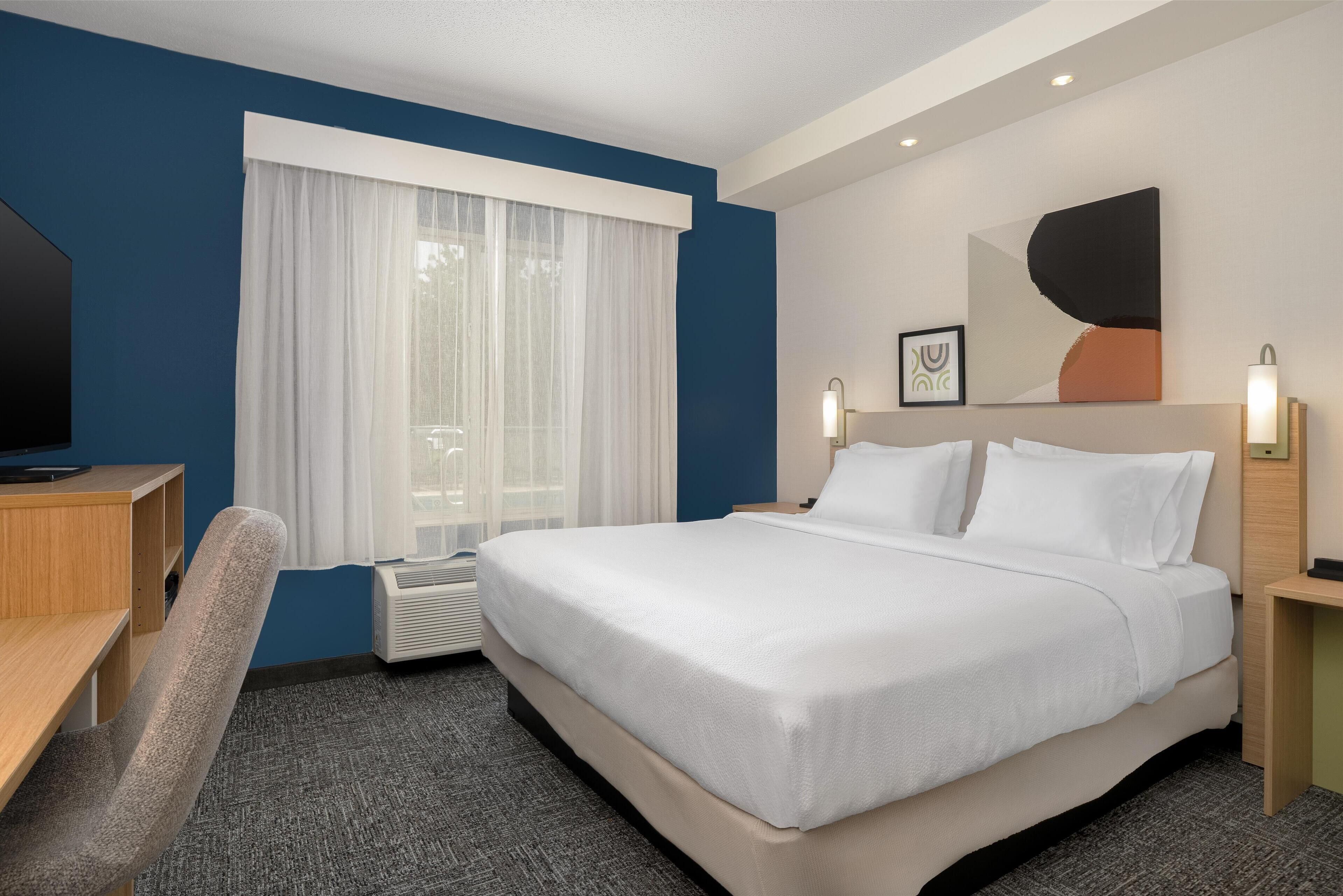 Een hotelkamer met een groot bed, een bureau, een stoel, een raam met gordijnen, een airconditioner en een kunstwerk aan de muur.