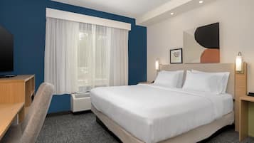 Quarto de hotel com uma cama grande, uma escrivaninha, uma cadeira, janela com cortinas, ar condicionado e uma obra de arte na parede.