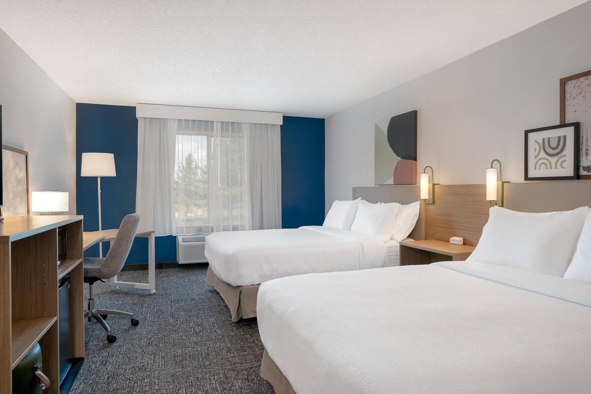 A hotel room with two beds, a desk, a chair, a lamp, and a window with curtains.