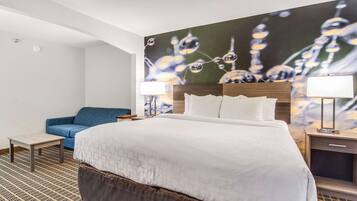 A hotel room with a large bed, a blue sofa, a small table, a nightstand with a lamp, and a wall with a floral painting.