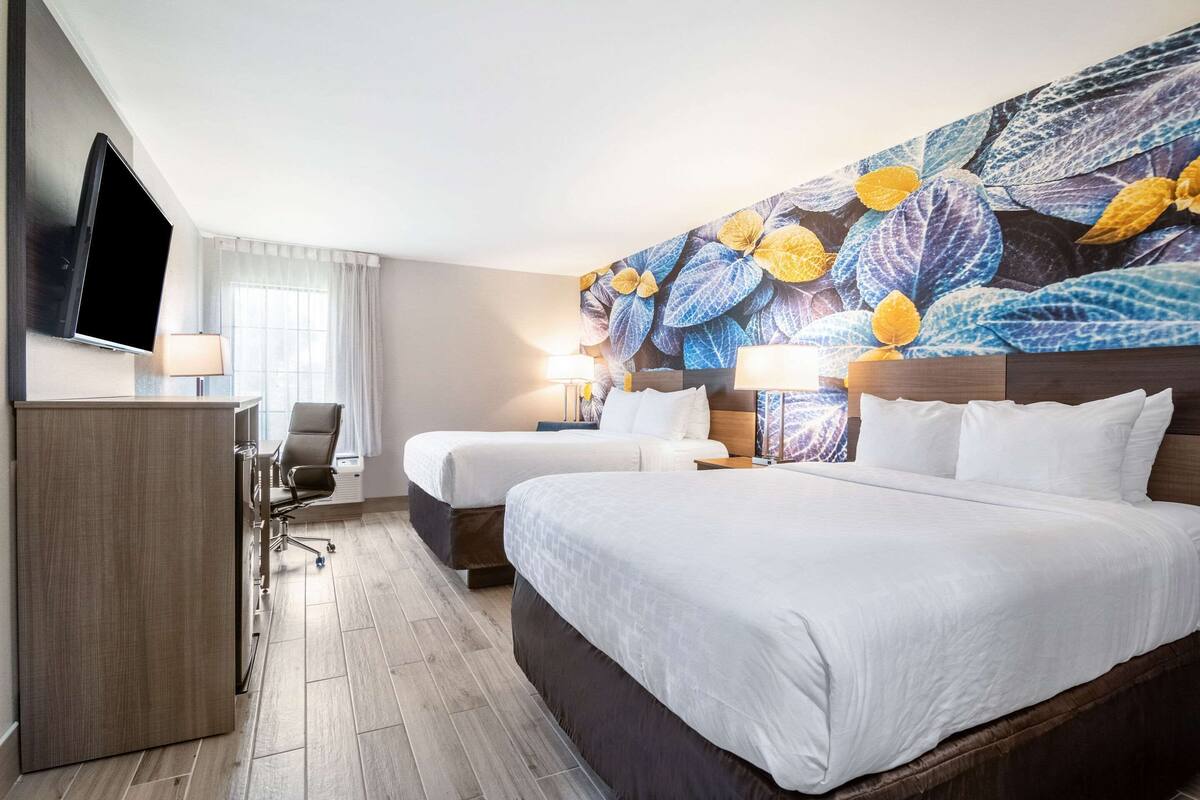 Habitación de hotel con dos camas, un escritorio, un televisor y un gran mural floral en la pared.