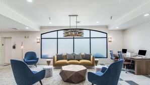 Un hall d’hôtel moderne avec un grand canapé, deux fauteuils bleus, une table basse hexagonale et un bureau équipé d’ordinateurs.