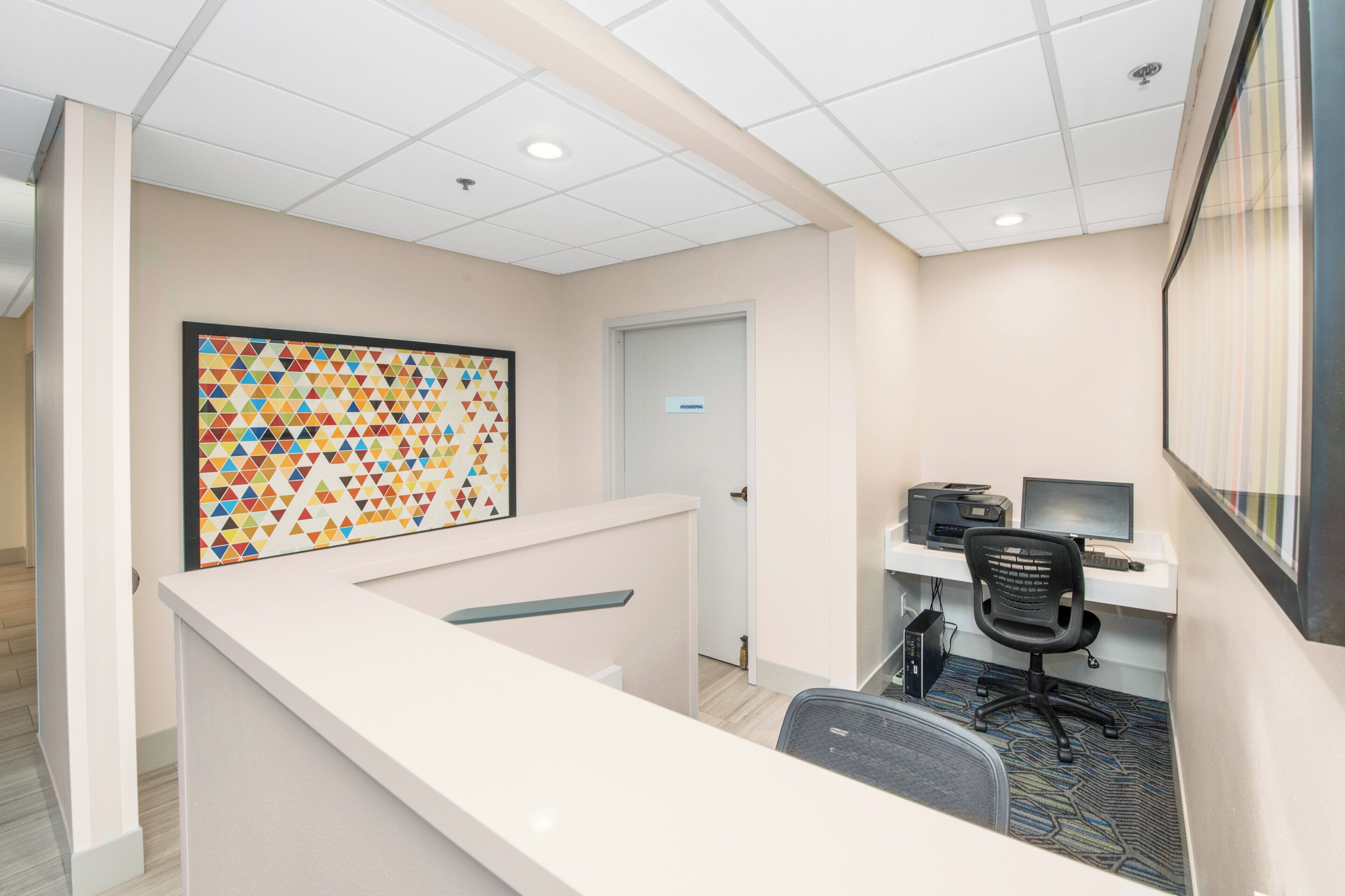A reception area with a white reception desk, a colorful geometric wall art, a closed door, and a computer desk with a chair.