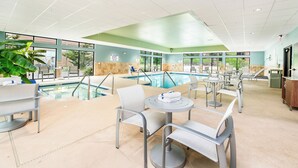 An indoor swimming pool area with multiple lounge chairs and tables, a pool with clear blue water, and a view of the outdoors through large windows. - Holiday Inn Express & Suites Delafield by IHG (Delafield)