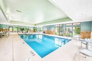 An indoor swimming pool with clear water, surrounded by lounge chairs and tables, and large windows offering a view of greenery outside.