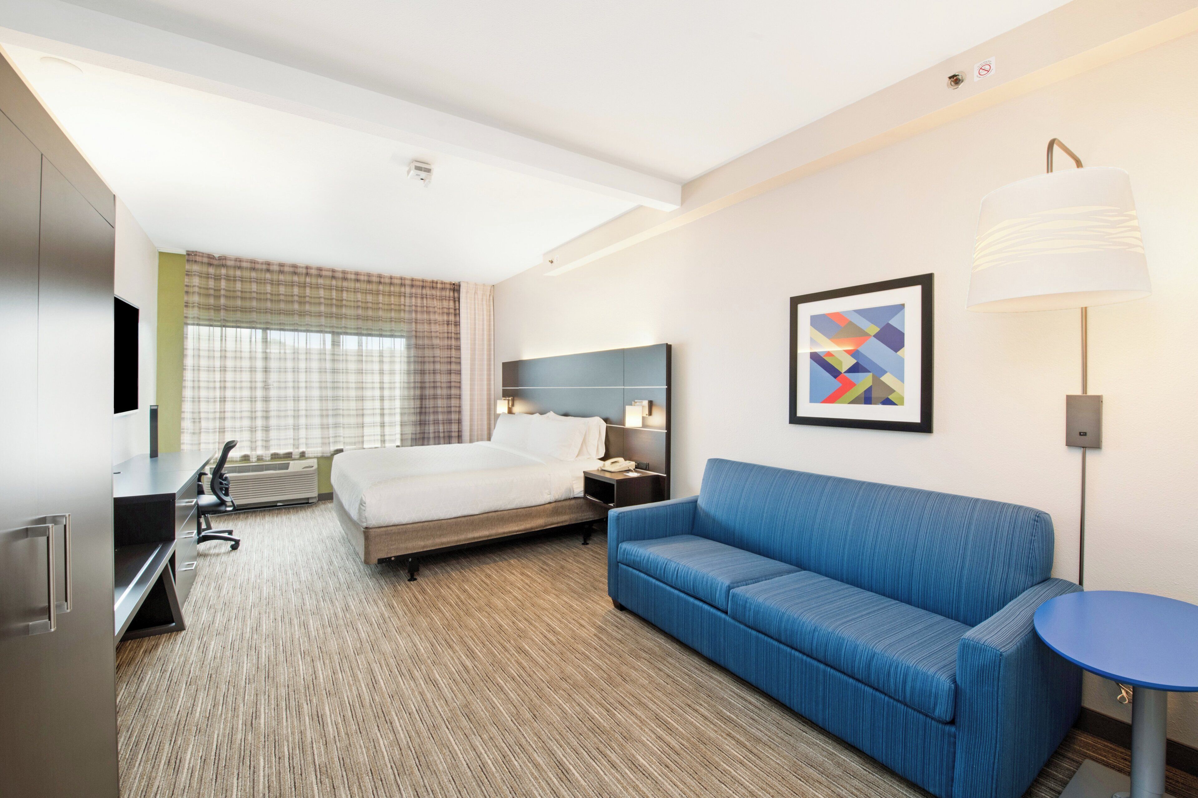 Una camera d'albergo con un letto, una scrivania, un divano blu, un tavolino, una lampada e un quadro appeso al muro.