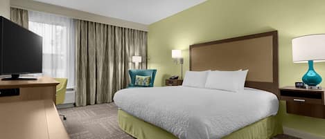 A hotel room with a large bed, a desk with a flat-screen TV, a chair, a nightstand with a lamp, and a window with curtains.