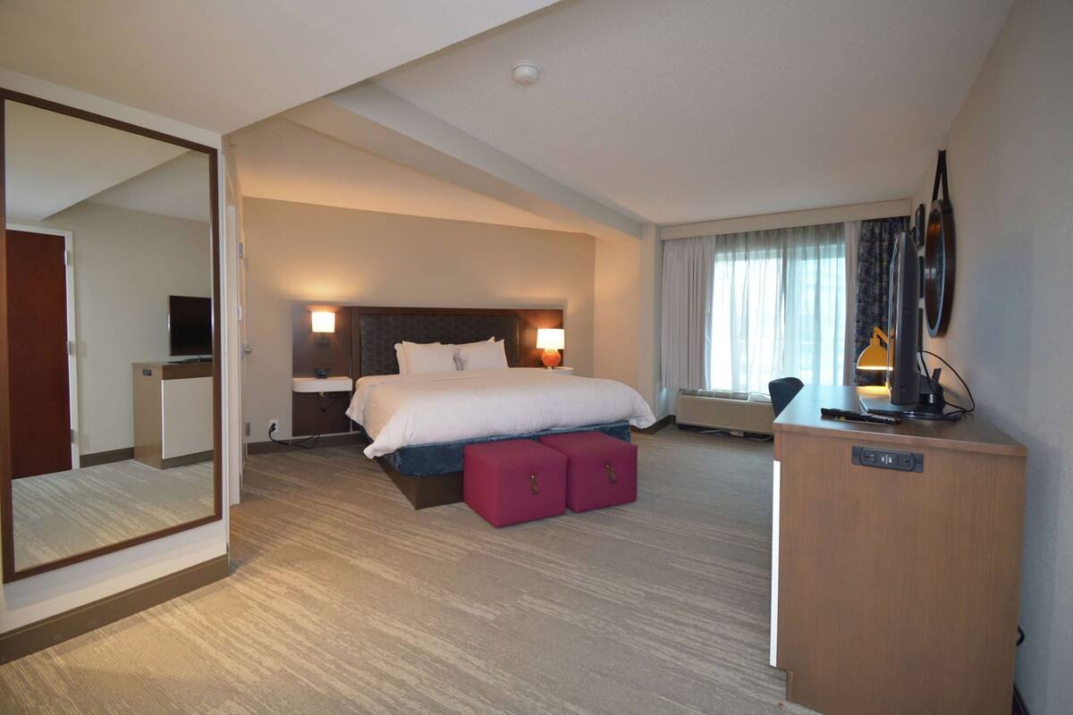A hotel room with a bed, bedside lamps, a desk with a computer, a mirror, and a window with curtains.
