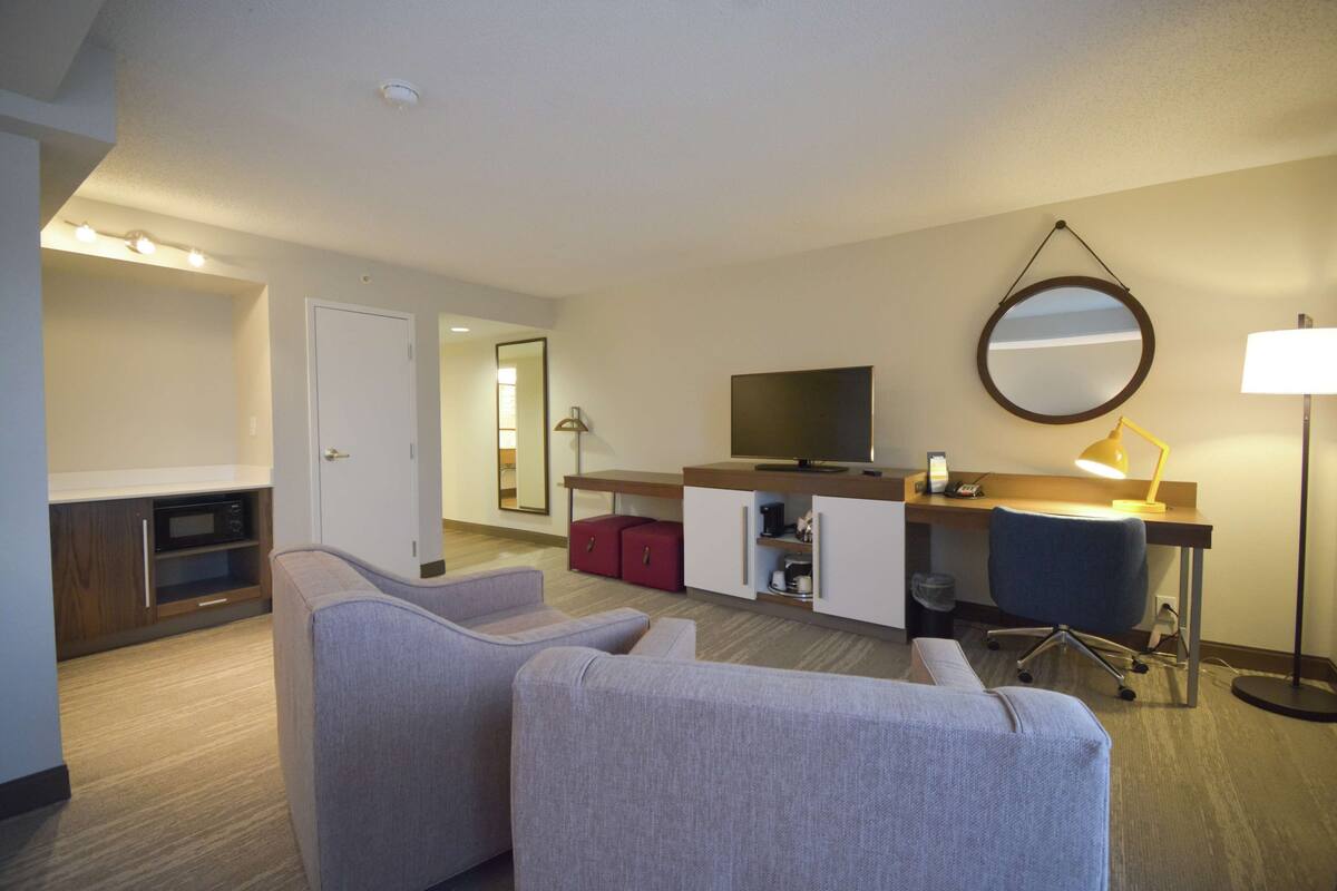 A hotel room with a kitchenette, a TV, a desk with a lamp, and two sofas.