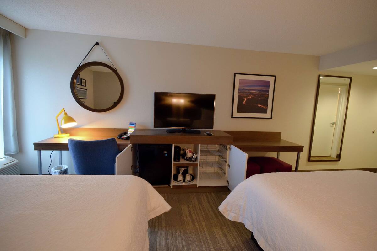 A hotel room with two beds, a desk with a computer, a TV, a desk lamp, a mirror, and a framed picture on the wall.