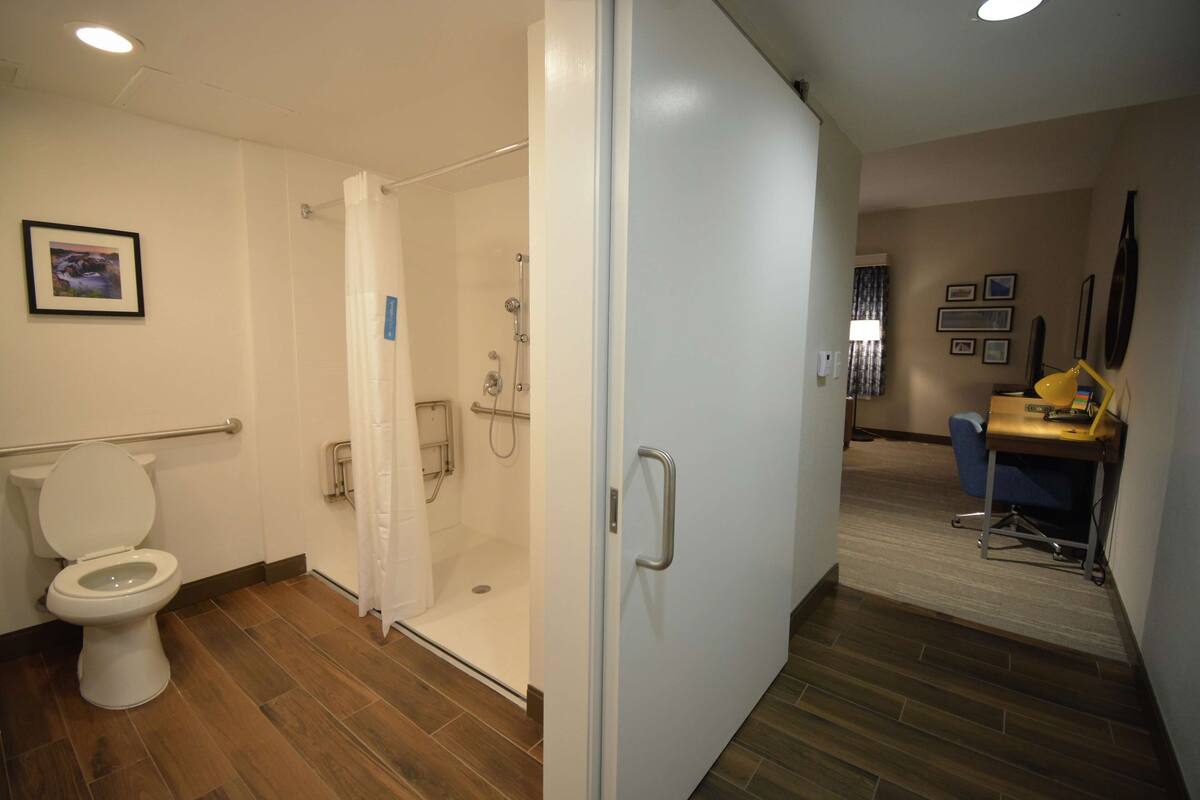 A bathroom with a toilet, shower, and a door leading to a room with a desk and chair.