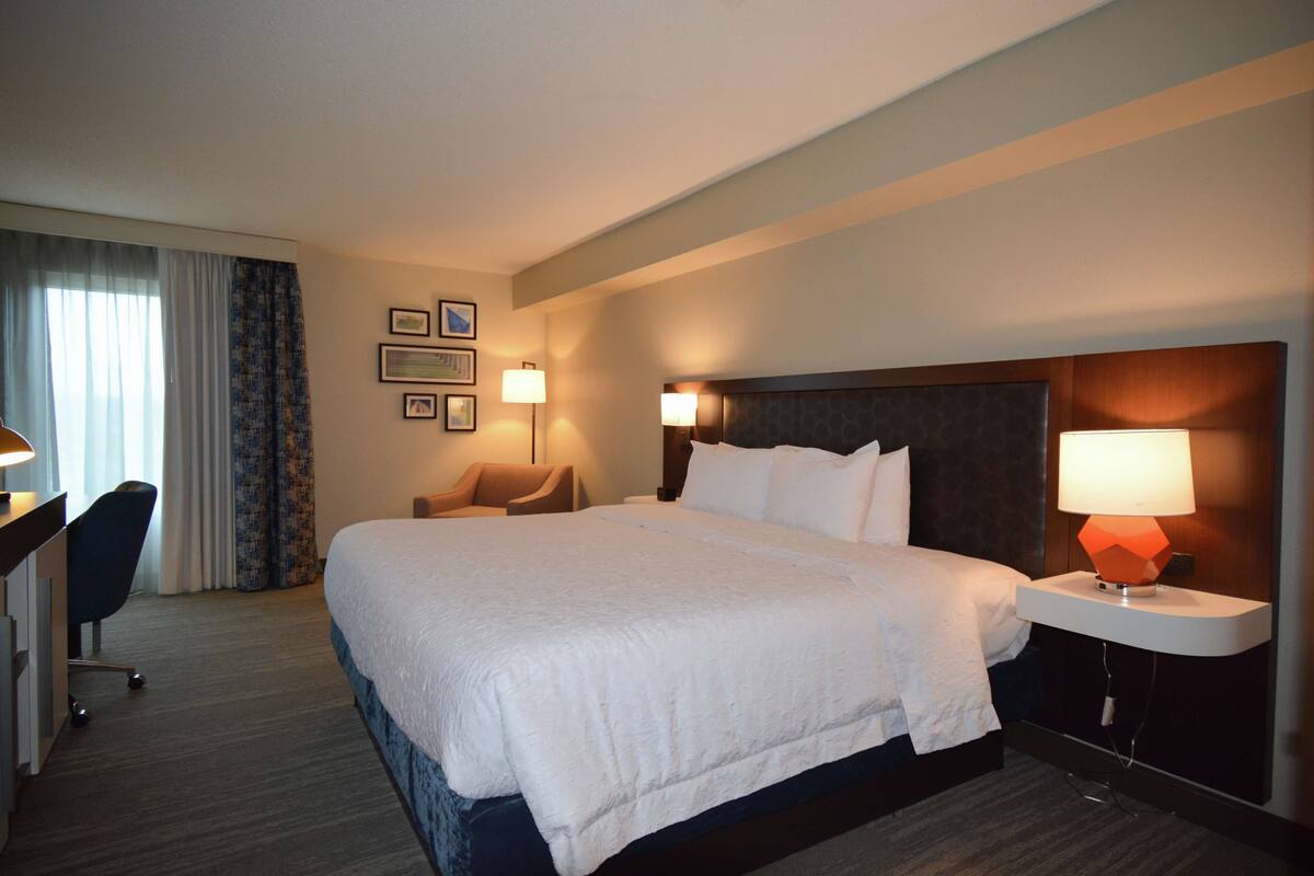 A hotel room with a large bed, a desk with a chair, two bedside lamps, and a headboard.