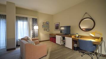 A hotel room with a sofa, a desk with a computer, a TV, a mirror, and a lamp.
