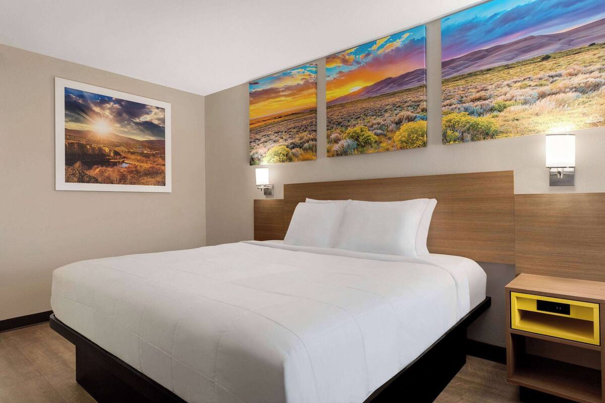 Ein Hotelzimmer mit einem großen Bett, einem Holz-Kopfteil, einem Nachttisch mit einer gelben Schublade und drei an der Wand hängenden Landschaftsbildern.