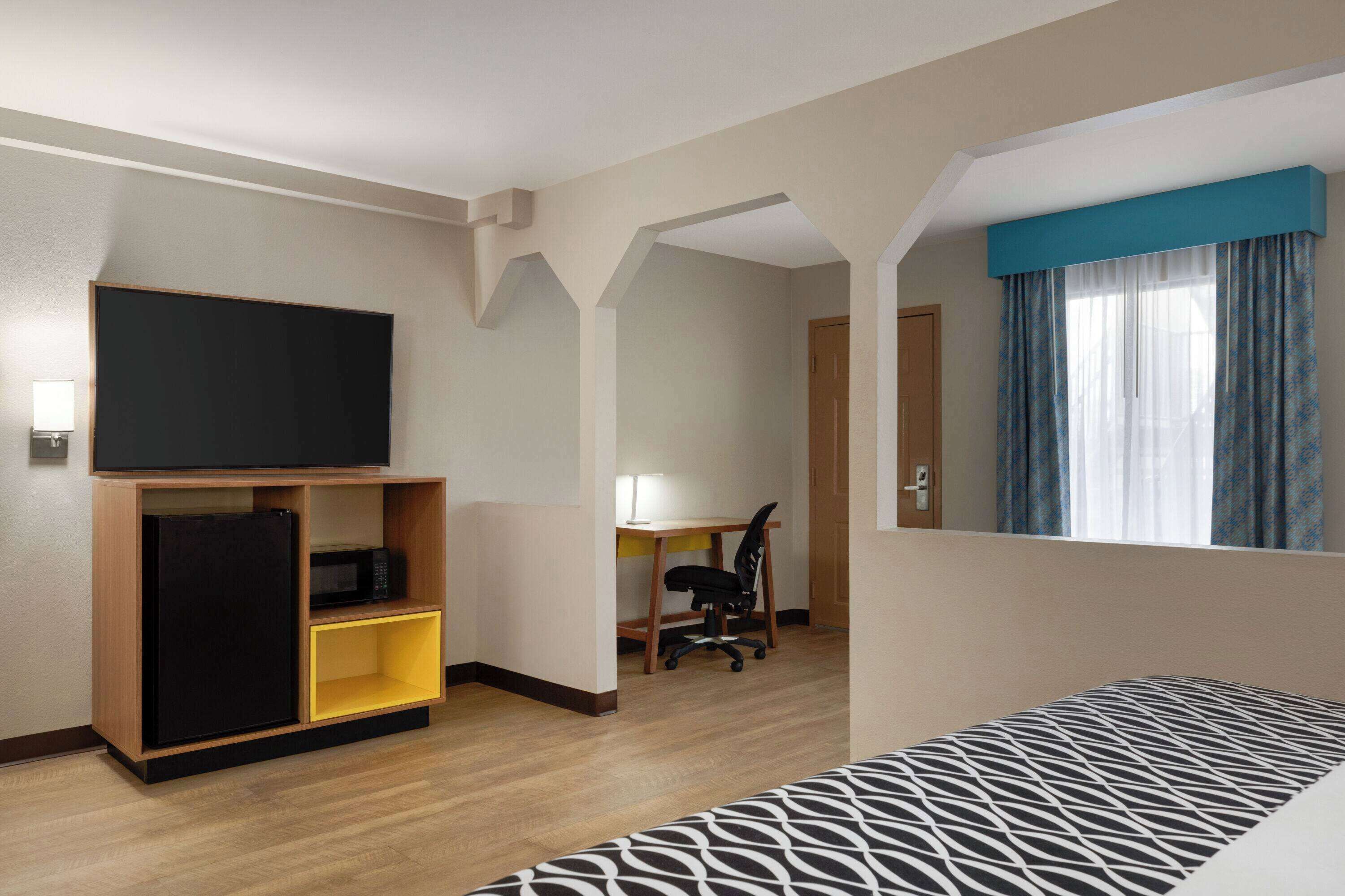 A hotel room with a bed, a TV on a stand, a desk with a chair, and a window with curtains.