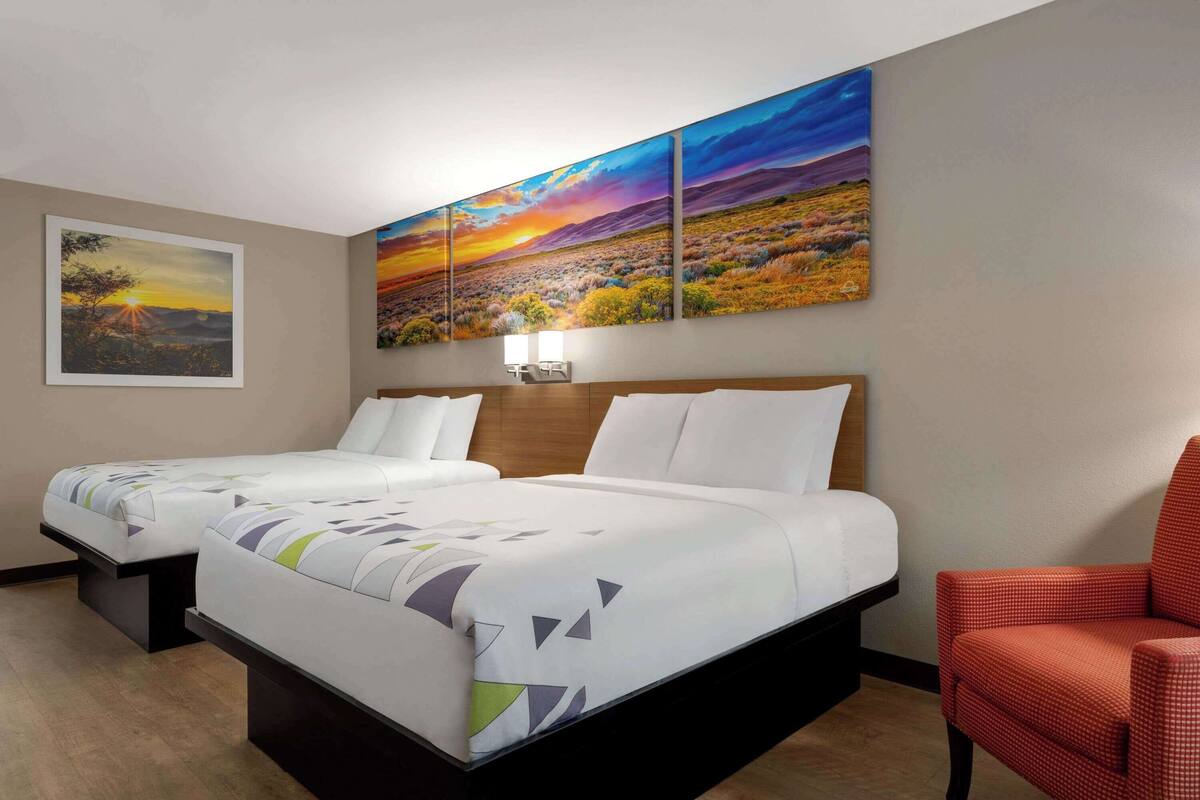 Ein Hotelzimmer mit zwei Betten, einem Nachttisch, einem roten Sessel und einem großen Landschaftsbild über den Betten.