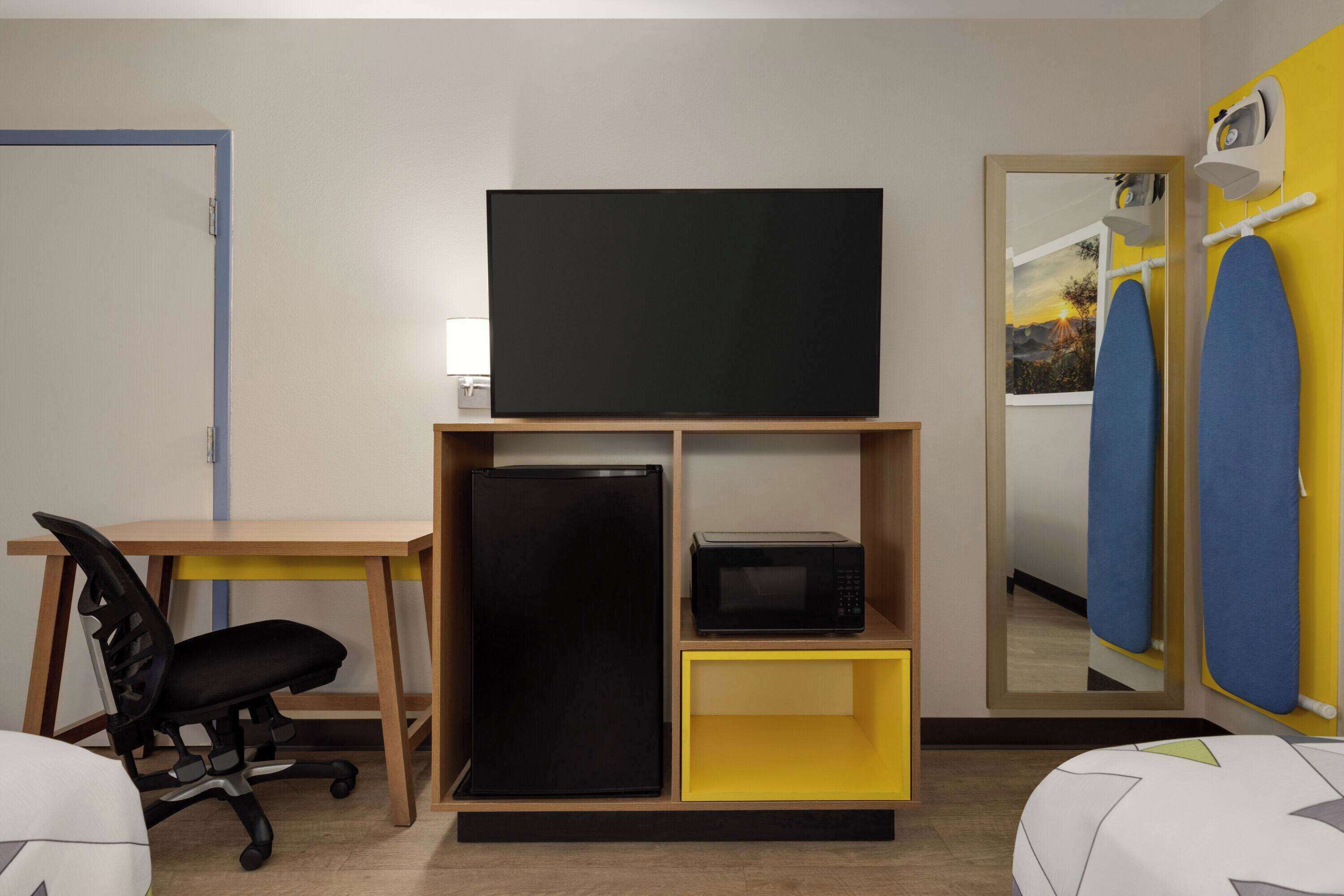 Una habitación con una televisión sobre un soporte de madera, un refrigerador negro, un microondas, un escritorio con una silla y una cama con cubrecolcha de diseño geométrico.
